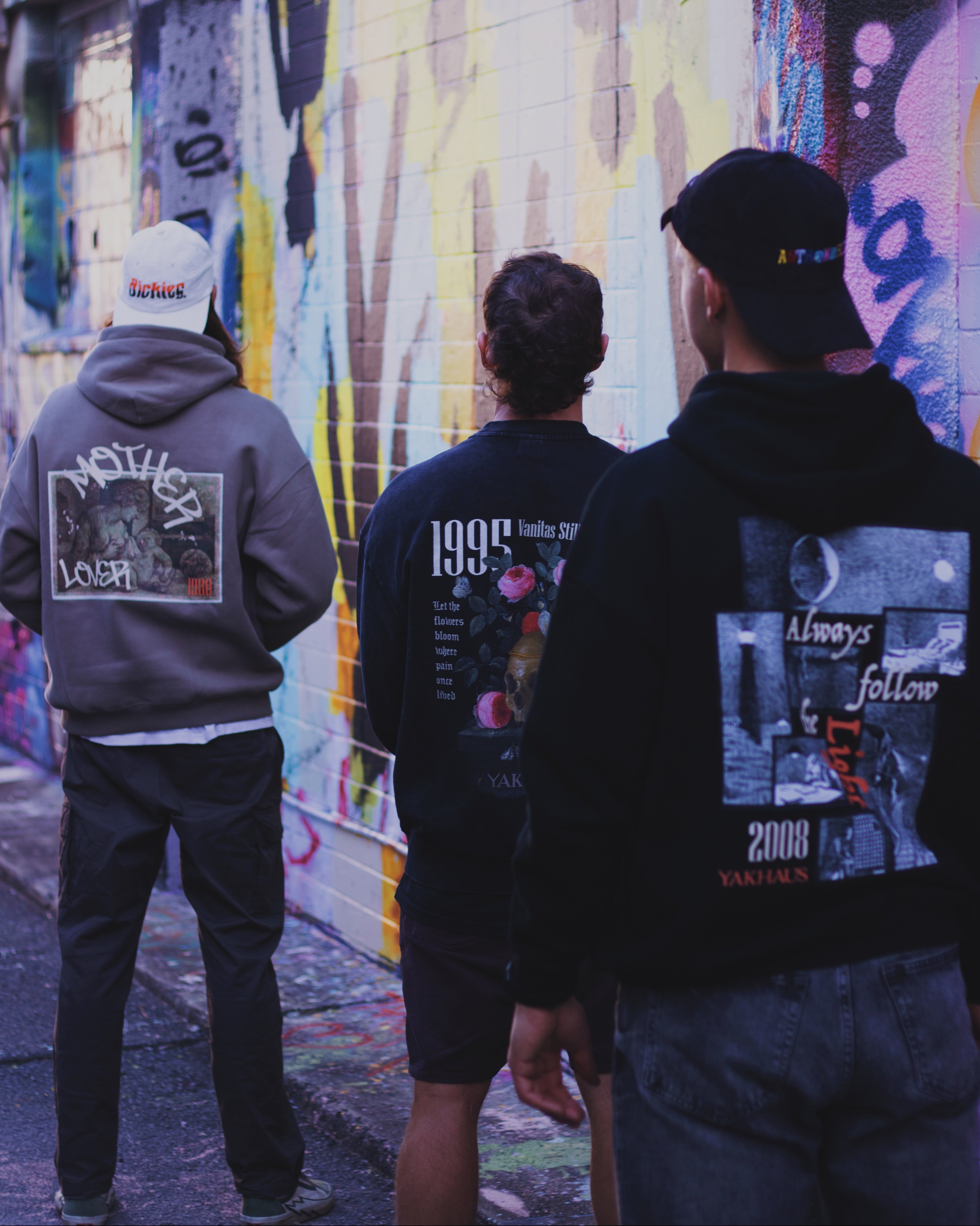 Three people wearing hoodies with graphic designs standing in front of a colorful mural.